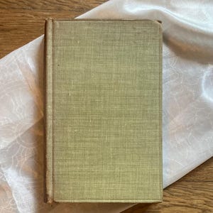 May include: A vintage book with a faded green cover. The book's spine and edges show signs of wear, suggesting its age. The cover has a textured appearance. The book rests on a white, patterned fabric, set against a wooden surface.