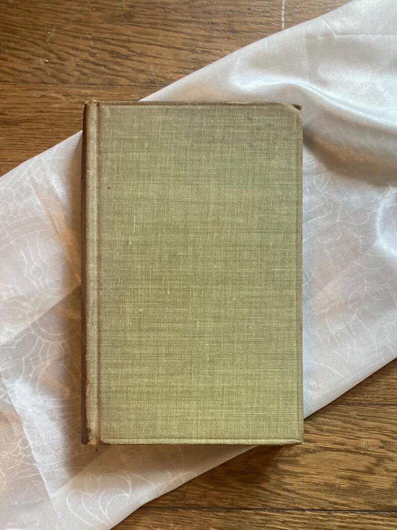 May include: A vintage book with a faded green cover. The book's spine and edges show signs of wear, suggesting its age. The cover has a textured appearance. The book rests on a white, patterned fabric, set against a wooden surface.