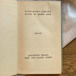 May include: Open book with aged, cream-colored pages. The text reads "Everyman's Library" and "Emerson's Essays, First and Second Series." The book's cover is a muted green. The book is open on a light-colored surface.