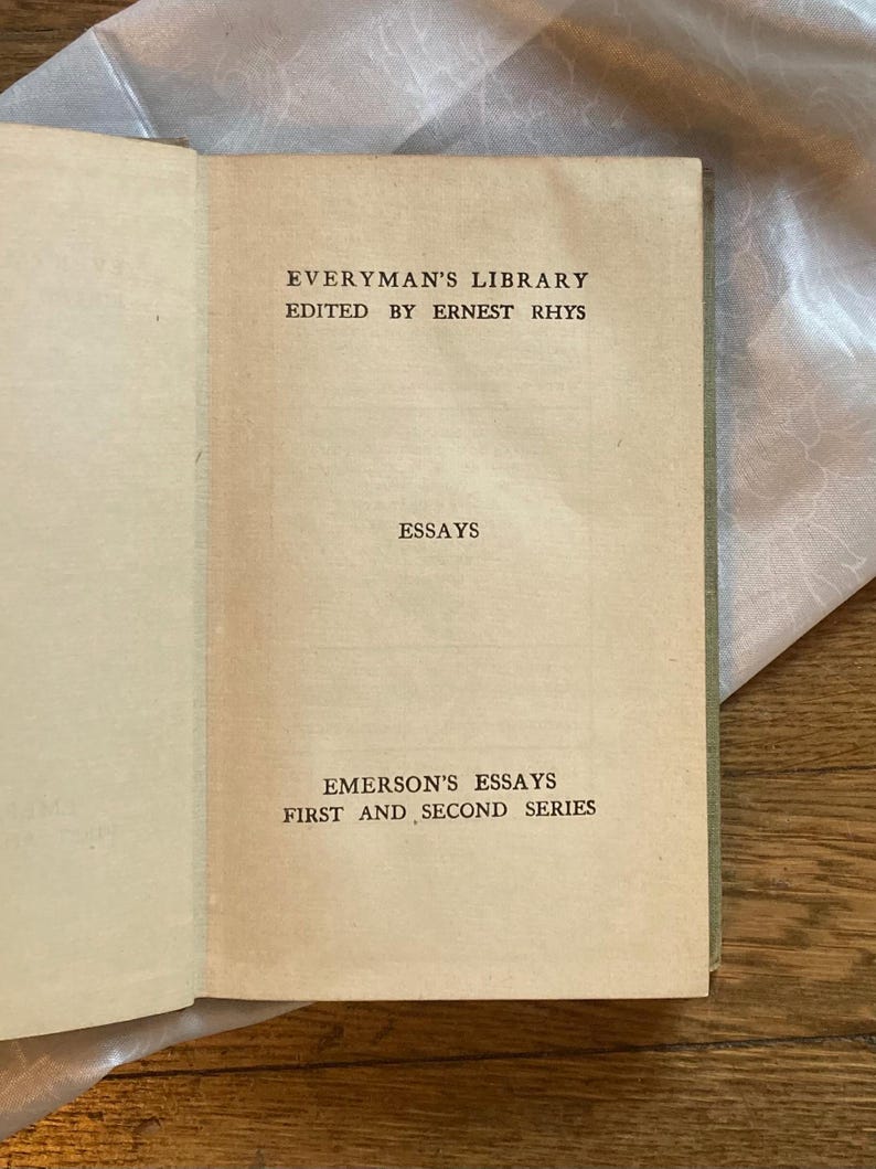 May include: Open book with aged, cream-colored pages. The text reads "Everyman's Library" and "Emerson's Essays, First and Second Series." The book's cover is a muted green. The book is open on a light-colored surface.