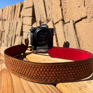 May include: A brown leather camera strap with a woven pattern and a red underside. A black Sony camera is in the background, resting on a wooden surface. The strap is designed to be worn around the neck.