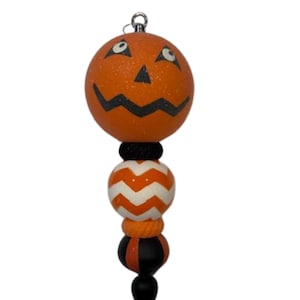 May include: A Halloween-themed decorative ornament featuring a smiling orange pumpkin head with black facial features. Below the pumpkin are decorative elements in orange, white, and black, culminating in a black, pointed base. The ornament is designed for seasonal decor.