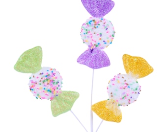 May include: A decorative candy pick featuring white candy-shaped ornaments with colorful sprinkles and glittery green, purple, and yellow accents. The candy is attached to a white stem, perfect for floral arrangements or craft projects.