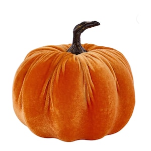 May include: A round, orange pumpkin made of soft velvet fabric with deep vertical pleats and folds. The pumpkin features a dark brown, textured, curved stem at the top center, providing a realistic contrast to the smooth, plush orange body.