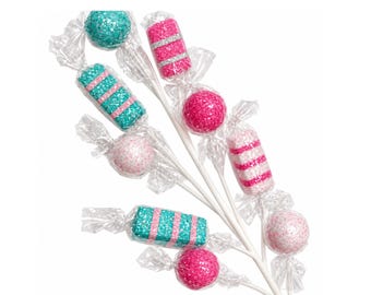 May include: A decorative spray featuring multiple candy-shaped ornaments on white sticks. The ornaments are wrapped in clear plastic and decorated with glitter in shades of teal, hot pink, and light pink, featuring striped and solid round patterns.