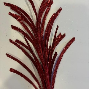 May include: A cluster of red glitter stems, possibly decorative, against a white background. The stems are curved and have a sparkling texture, suggesting a festive or craft-related item.