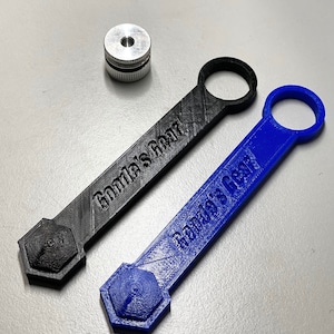 May include: A top-down shot features two 3D-printed tools, one black and one blue, each with a loop, a rectangular handle with the text "Gonda's Gear", and a hexagonal end. A small, silver metal cylinder sits nearby.