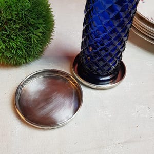 May include: A cobalt blue glass vase with a diamond pattern, resting on a silver-colored base. A small, round, silver-colored tray sits nearby. A green, spherical plant is in the background.