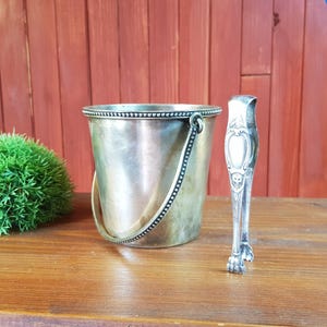 May include: A vintage silver ice bucket with a handle and a pair of ornate ice tongs. The bucket is cylindrical with a beaded rim and handle. The tongs have a decorative design. The items are on a wooden table.
