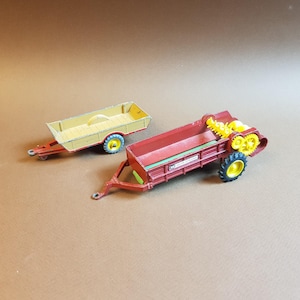 May include: Two toy farm vehicles: a red and yellow manure spreader and a yellow trailer. The manure spreader has yellow gears and black and yellow wheels. The trailer has a yellow bed and blue and yellow wheels.