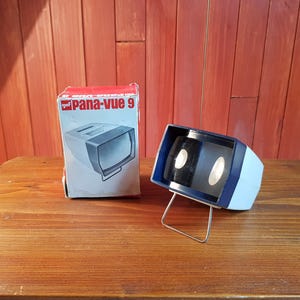 Peut inclure: Un ancien lecteur Pana-Vue 9 et sa boîte d'origine. Le lecteur est bleu clair et noir, avec un support en fil d'argent. La boîte est blanche et rouge, avec un graphique du lecteur. Les articles sont sur une surface en bois.