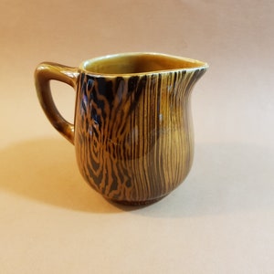 May include: A small, brown ceramic pitcher with a handle and a spout. The pitcher has a wood grain pattern in shades of brown and black. The interior is a lighter brown color.