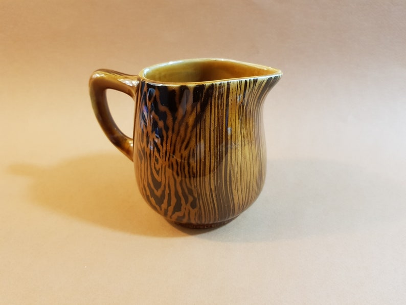 May include: A small, brown ceramic pitcher with a handle and a spout. The pitcher has a wood grain pattern in shades of brown and black. The interior is a lighter brown color.