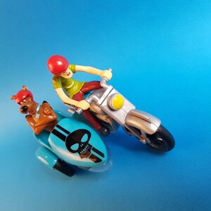 May include: A blue and silver toy motorcycle with a skull design on the sidecar. A cartoon character with a red helmet is riding the motorcycle and a brown dog character is in the sidecar.