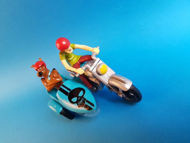 May include: A blue and silver toy motorcycle with a skull design on the sidecar. A cartoon character with a red helmet is riding the motorcycle and a brown dog character is in the sidecar.
