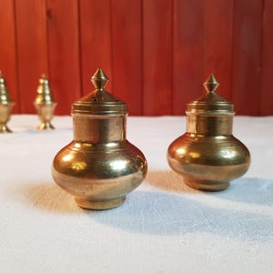 May include: Two antique brass salt and pepper shakers with a bulbous body and a conical lid. The shakers have a warm, golden hue and are placed on a white tablecloth. In the background, there are two additional brass containers.