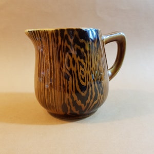 May include: A brown ceramic pitcher with a wood grain pattern. The pitcher has a handle and a spout for pouring. The wood grain design features dark brown and black vertical lines. The pitcher is likely used for serving beverages.