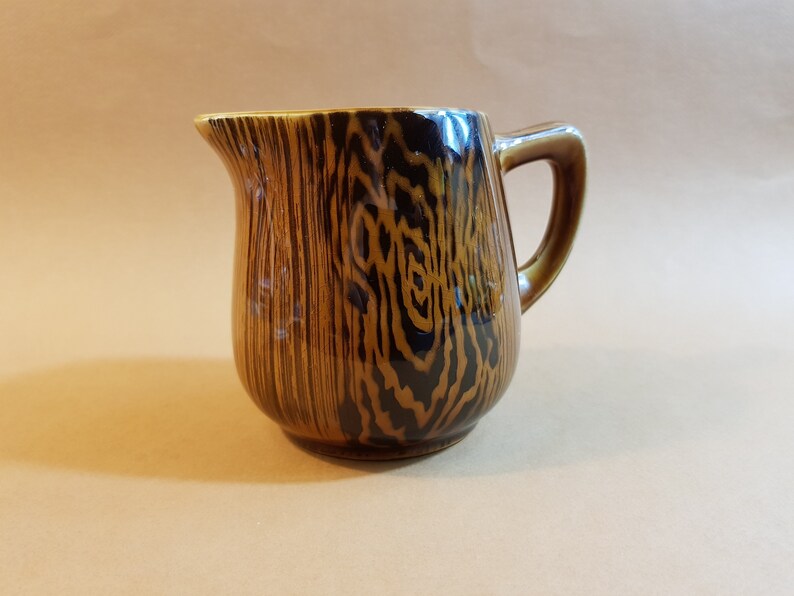 May include: A brown ceramic pitcher with a wood grain pattern. The pitcher has a handle and a spout for pouring. The wood grain design features dark brown and black vertical lines. The pitcher is likely used for serving beverages.
