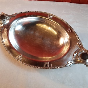 Puede incluir: Bandeja ovalada plateada con asas ornamentadas y un borde decorativo. La bandeja tiene una superficie pulida y reflectante y presenta diseños florales y geométricos estilizados. Las asas tienen detalles circulares oscuros.