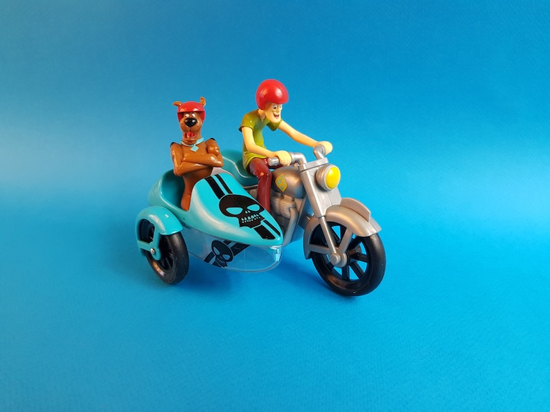 May include: A blue and silver toy motorcycle with a sidecar. The motorcycle has a skull and crossbones design on the sidecar. A cartoon character is riding the motorcycle and another cartoon character is sitting in the sidecar.