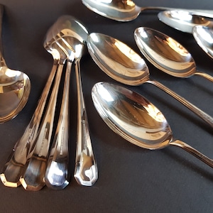 Puede incluir: Conjunto de cucharas de plata pulida dispuestas sobre una superficie oscura. Las cucharas tienen mangos alargados y cuencos ovalados. Algunas están agrupadas, otras separadas, mostrando sus superficies reflectantes.