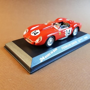 May include: A red 1956 Ferrari 250TR model car with a white number 14 on the side. The car is on a black display base with the text "24 Heures du Mans" and "Ferrari 250TR" printed on it.