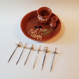 Puede incluir: Un juego de servir aceitunas de terracota con un plato decorativo, una taza pequeña y un jarrón. El plato tiene la palabra "Olives" y un diseño. Seis pinchos de aceitunas plateados con forma de espada están en primer plano.