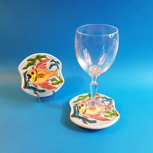 Könnte beinhalten: Ein klares Weinglas steht auf einem Keramikuntersetzer mit handgemaltem Fischdesign. Der Untersetzer ist hellblau mit einem gelben, roten und grünen Fisch. Ein weiterer Untersetzer mit dem gleichen Design wird auf einem Metallständer präsentiert.