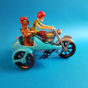 May include: A blue and silver toy motorcycle with a sidecar. A brown cartoon dog with a green shirt is sitting in the sidecar. A red-headed cartoon character is driving the motorcycle.