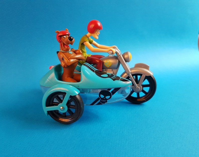 May include: A blue and silver toy motorcycle with a sidecar. A brown cartoon dog with a green shirt is sitting in the sidecar. A red-headed cartoon character is driving the motorcycle.