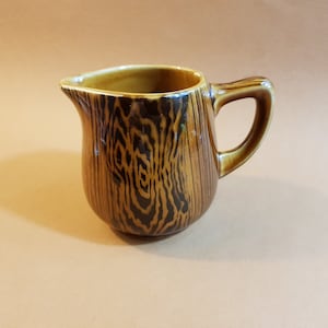 Puede incluir: Una jarra de cerámica con diseño de vetas de madera. La jarra es marrón con detalles marrón oscuro y un asa. Tiene un pico vertedor. Es un artículo de cocina decorativo.