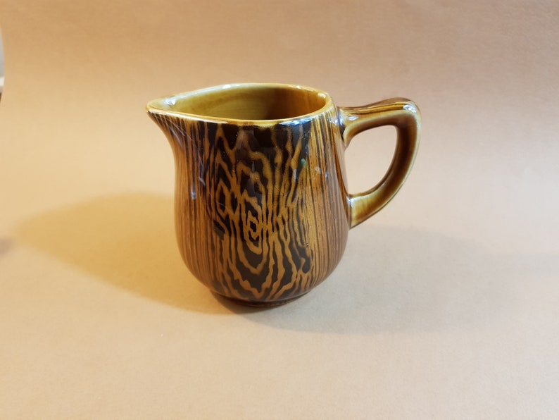 May include: A ceramic creamer with a wood grain design. The creamer is brown with dark brown accents and a handle. The creamer has a spout for easy pouring. The creamer is a decorative kitchen item.