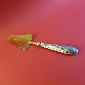 May include: A gold and silver cake server with an ornate handle. The blade is gold and the handle is silver with a floral design.