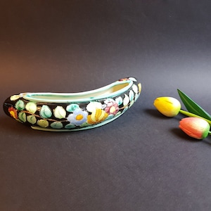Könnte beinhalten: Ein schwarzer Keramik-Blumentopf in Bootsform, verziert mit bunten Blumenmustern und grünen Blättern. Der Innenraum ist weiß mit einem türkisfarbenen Rand. Zwei künstliche Tulpen liegen neben dem Topf.