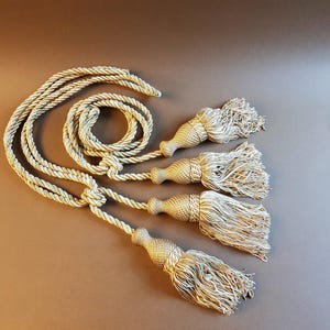 May include: Four gold tassel tiebacks with a rope design. The tassels are made of a soft, fluffy material and are attached to a thick, braided rope.