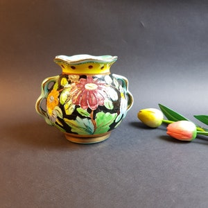 Könnte beinhalten: Eine farbenfrohe Keramikvase mit floralem Design. Die Vase hat einen dunklen Hintergrund mit roten, gelben und blauen Blumen. Sie hat zwei Henkel und einen gewellten Rand. Die Vase ist etwa 15 cm hoch.