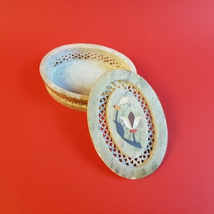 May include: A carved oval-shaped box made of light gray stone with a floral design on the lid. The lid is open and the inside of the box is visible. The box is on a red background.