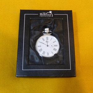May include: A silver pocket watch with Roman numeral markings on a white face, in a black box with "The Heritage Collection" printed on the top.