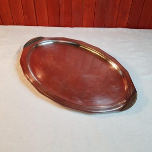 May include: An oval serving tray with a polished, reddish-brown surface and a silver-colored rim. The tray has dark brown wooden handles on each end. The tray is on a white tablecloth.