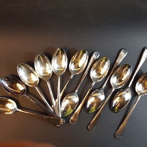 May include: A collection of silver spoons arranged on a dark surface. The spoons have oval bowls and long, slender handles. The spoons are reflective, showing the surrounding environment.