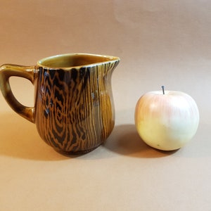 May include: A brown ceramic pitcher with a wood grain pattern and a handle. Next to the pitcher is a realistic-looking apple with a pink and yellow gradient. The background is a solid tan color.