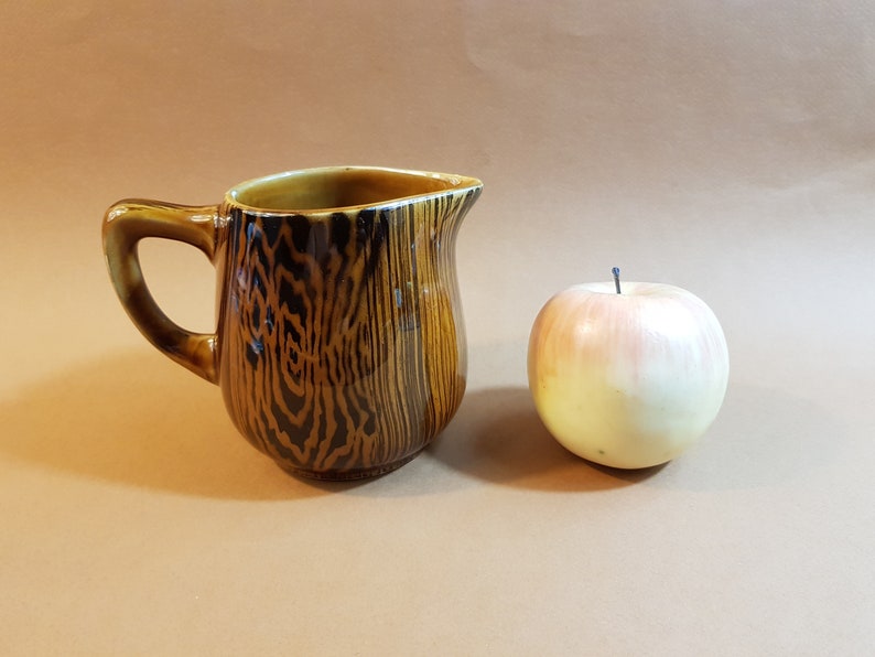 May include: A brown ceramic pitcher with a wood grain pattern and a handle. Next to the pitcher is a realistic-looking apple with a pink and yellow gradient. The background is a solid tan color.