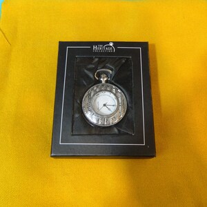 May include: A silver pocket watch with Roman numeral markings around the face, in a black box with "The Heritage Collection" printed on the top.