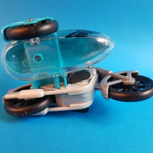 May include: A turquoise and silver toy motorcycle with a clear plastic canopy. The motorcycle has three wheels and the text "The Gizmo" is printed on the side.