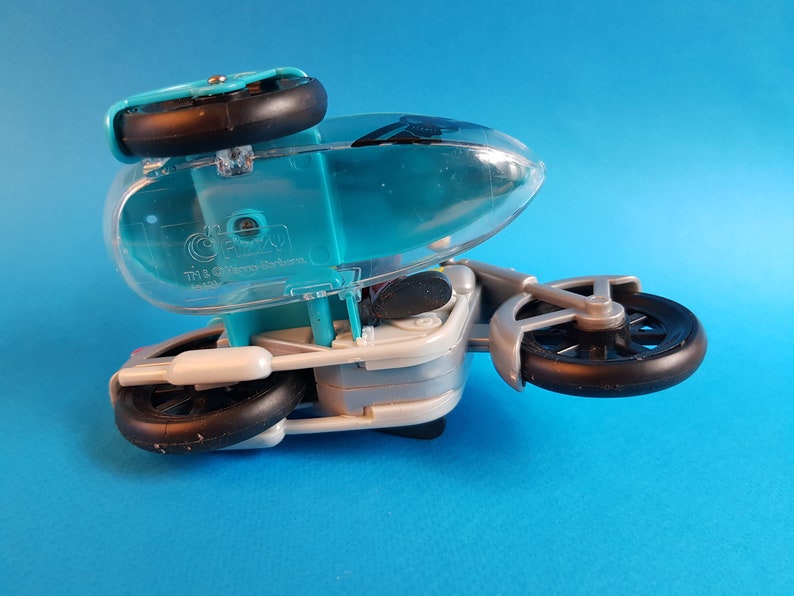 May include: A turquoise and silver toy motorcycle with a clear plastic canopy. The motorcycle has three wheels and the text "The Gizmo" is printed on the side.