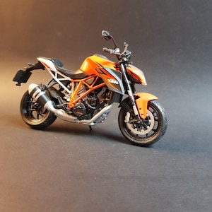 May include: Orange and black KTM motorcycle model with silver and white accents. The motorcycle has a detailed engine, wheels, and handlebars. The model is set against a dark background.