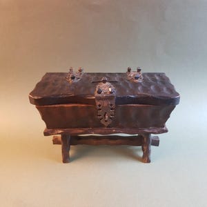 May include: A small wooden box with a hinged lid and a metal latch. The box is sitting on a wooden stand with four legs. The box is dark brown and has a carved design on the lid.