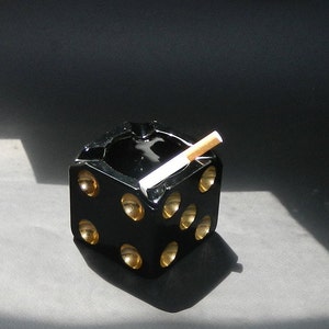 May include: Black glass ashtray shaped like a dice with gold accents. A cigarette is resting in the ashtray.