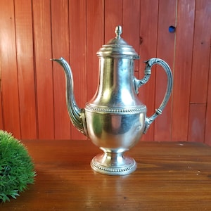 May include: A silver-colored coffee pot with a lid, curved spout, and ornate handle. The pot sits on a round base and is displayed on a wooden surface. The background is a red wooden wall.