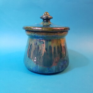 May include: A ceramic jar with a lid, featuring a green, blue, and brown iridescent glaze. The jar has a rounded body and a domed lid with a knob.